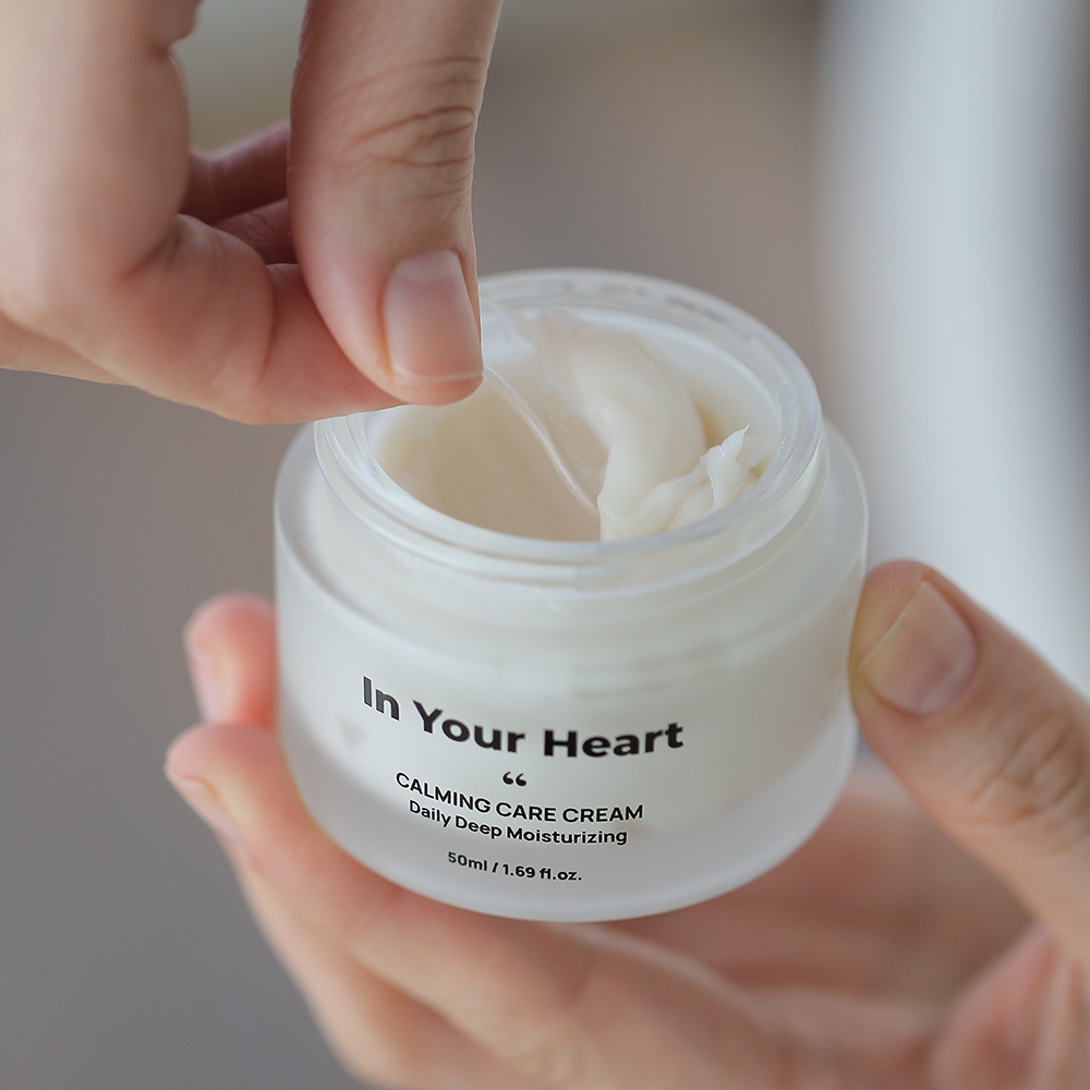 A hand scooping a creamy texture of the 'In Your Heart Calming Care Cream' from its jar, showcasing the smooth, rich formula.