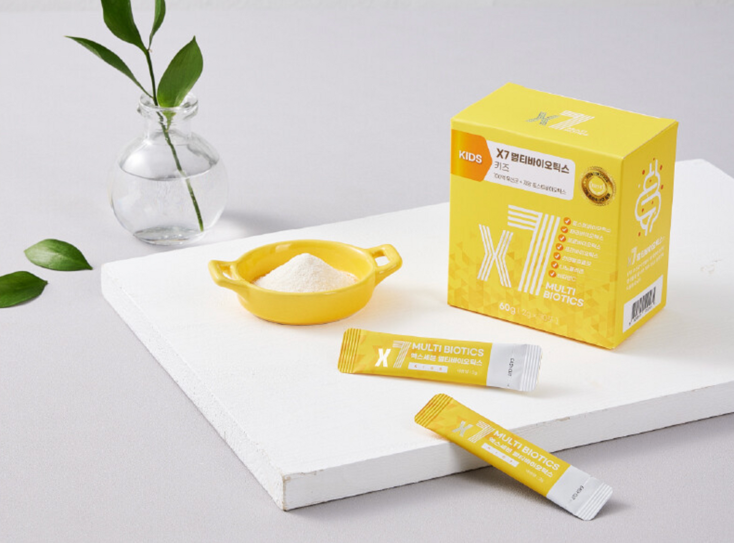 A yellow box of X7 Kids Multi Biotics with two stick sachets in front, accompanied by a small bowl of powdered supplement, a clear vase with greenery, and a clean white surface.