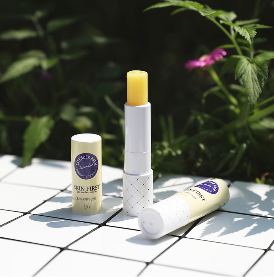 A close-up of an open tube of [DressingTable] Skin First Lavender Soothing Balm Mini resting outdoors on a tiled surface surrounded by greenery. The yellow balm is extended, and its elegant design is complemented by the fresh garden setting.