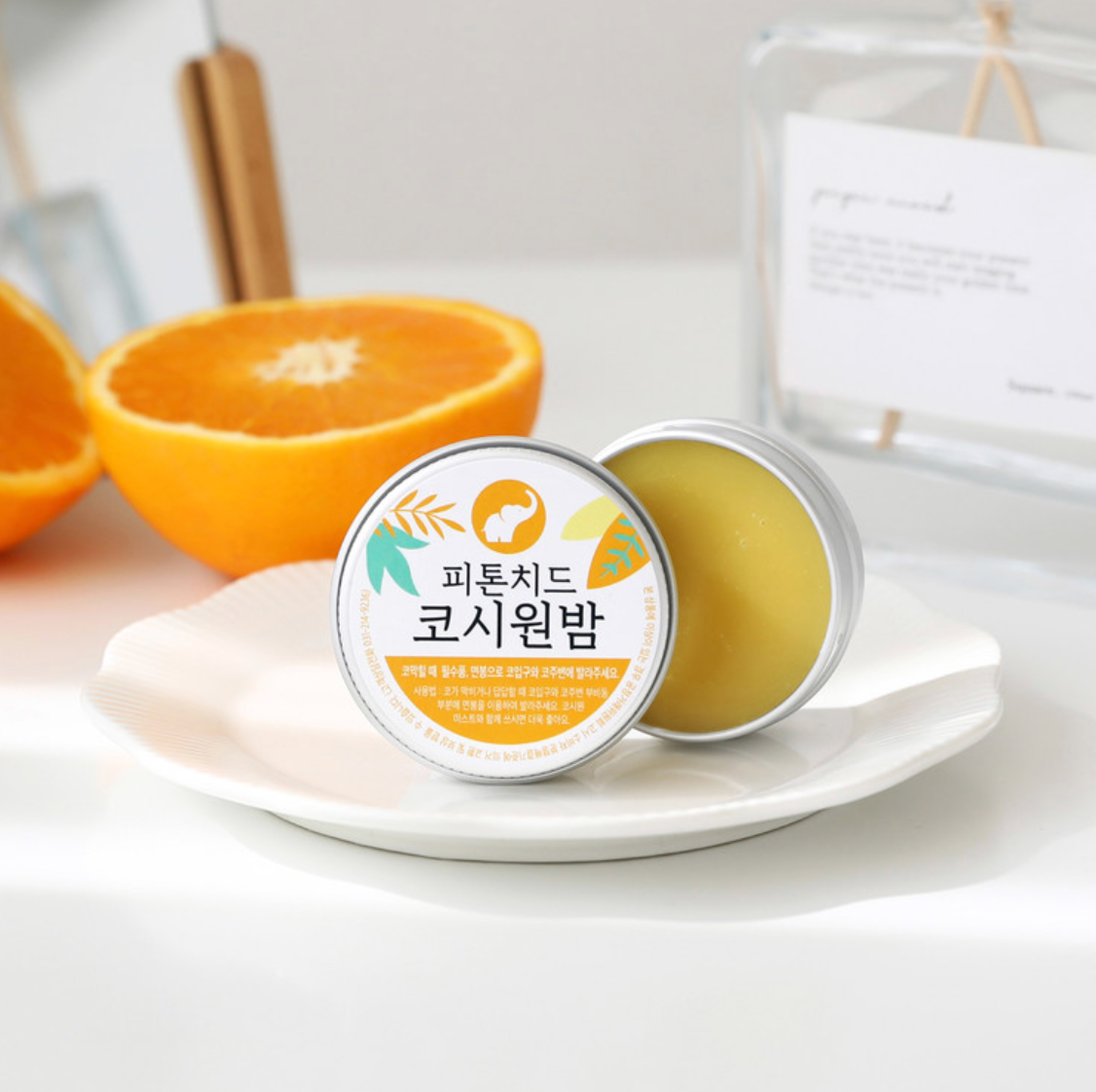 tin can of nose comfort balm with lid open to show orange balm, and a real orange cut open behind it.