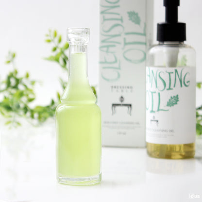 A close-up image of a transparent glass bottle filled with light green oil, elegantly displayed with a blurred background of green foliage.