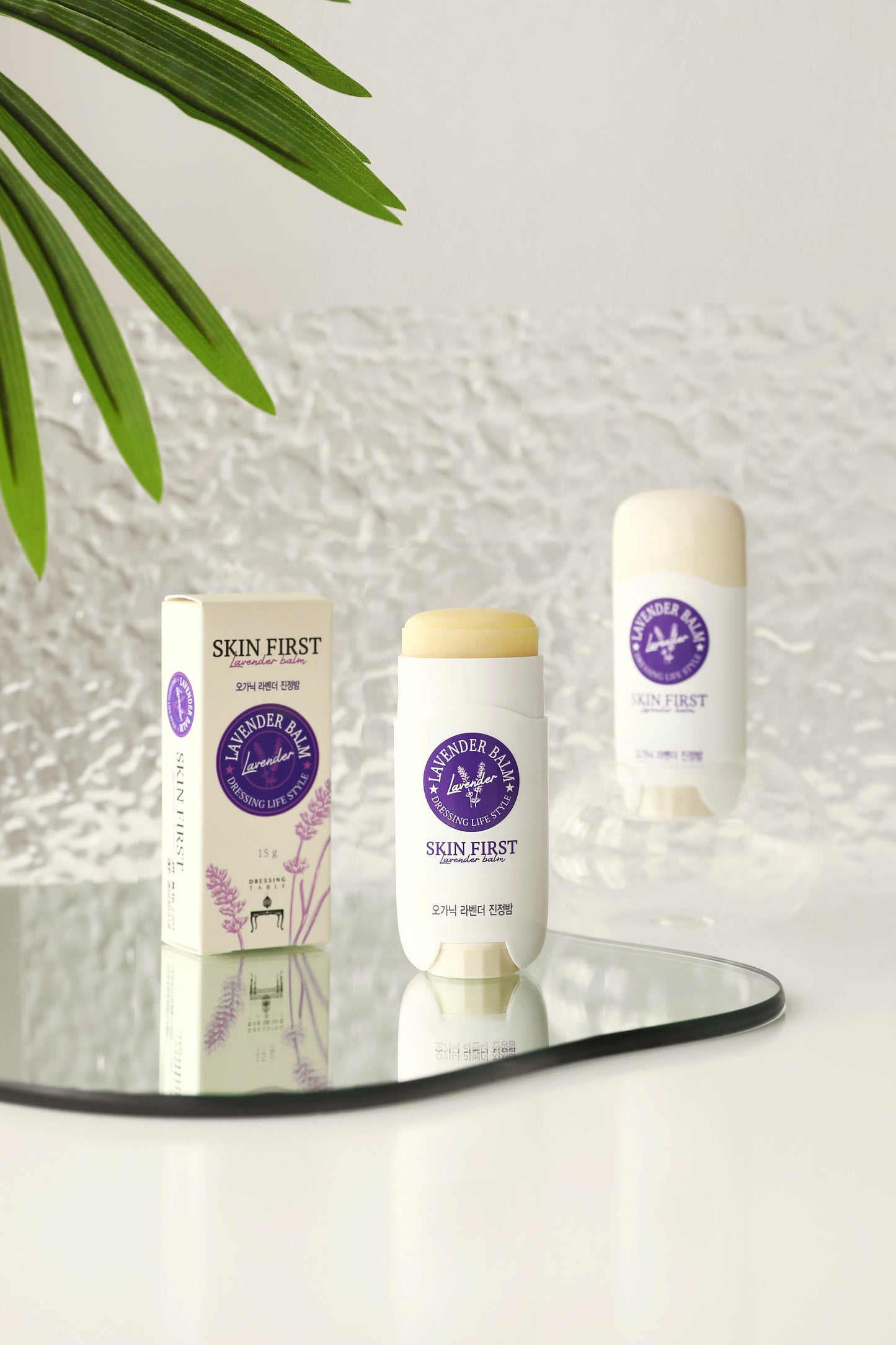 DressingTable Skin First Lavender Soothing Balm Stick placed on a reflective surface with its packaging in the background, showcasing the sleek, travel-friendly design and soft cream color.