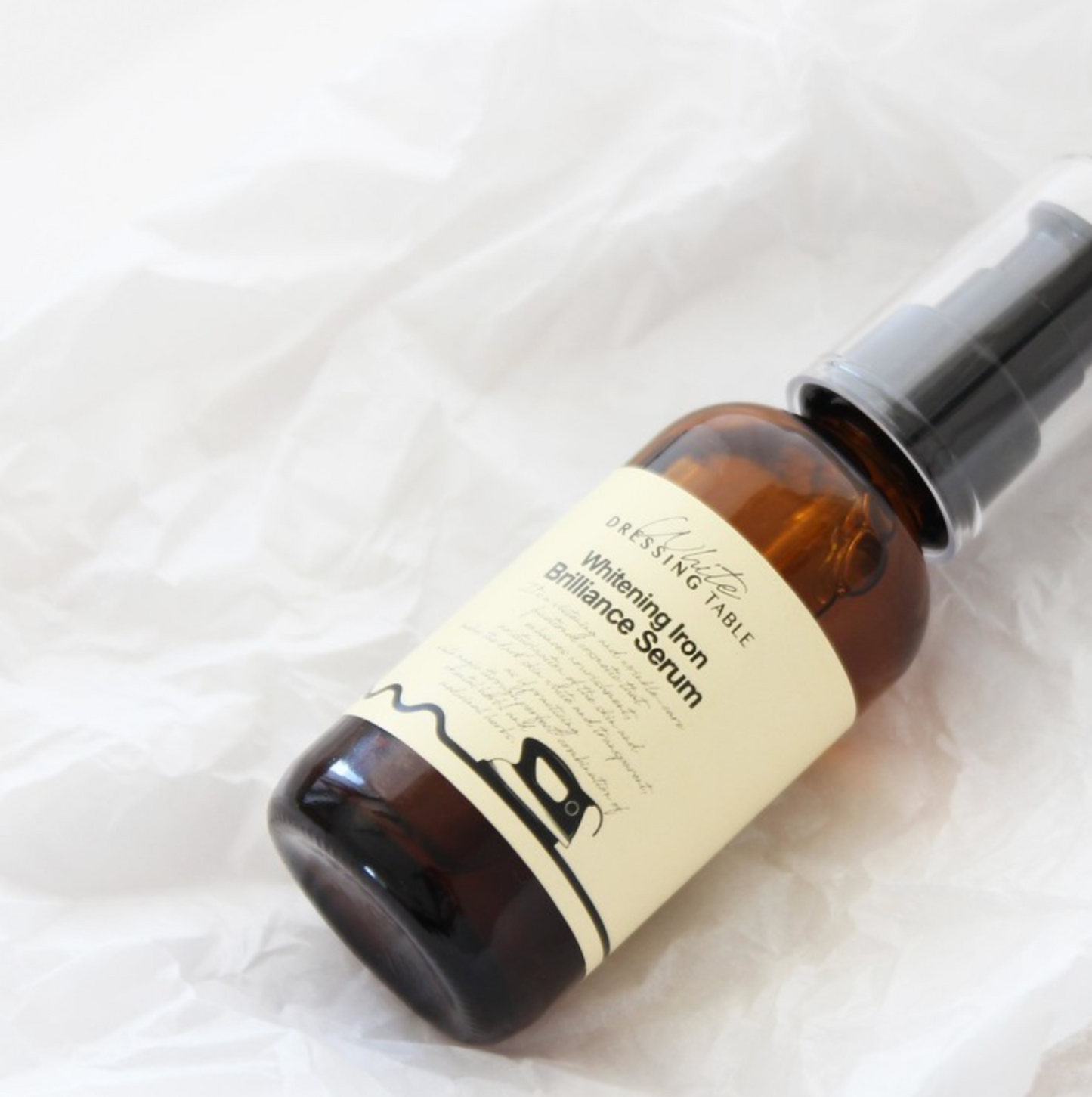 Brightening Iron Brilliance Serum lying on crinkled white paper, highlighting its amber glass bottle and minimalist label design. The soft texture of the paper enhances the luxurious feel of the product.