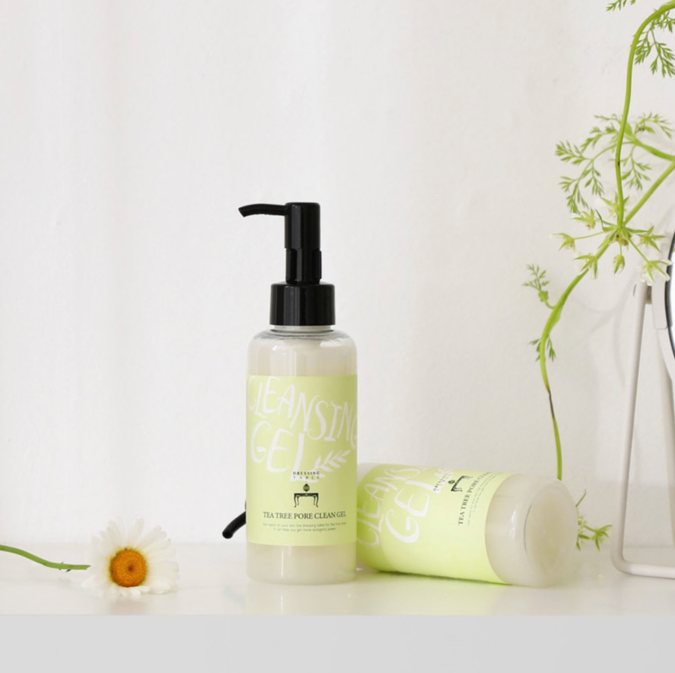 Two bottles of [DressingTable] Tea Tree Pore Cleansing Gel, one standing upright and the other lying on its side, displayed with greenery and a white daisy on a clean, bright surface.