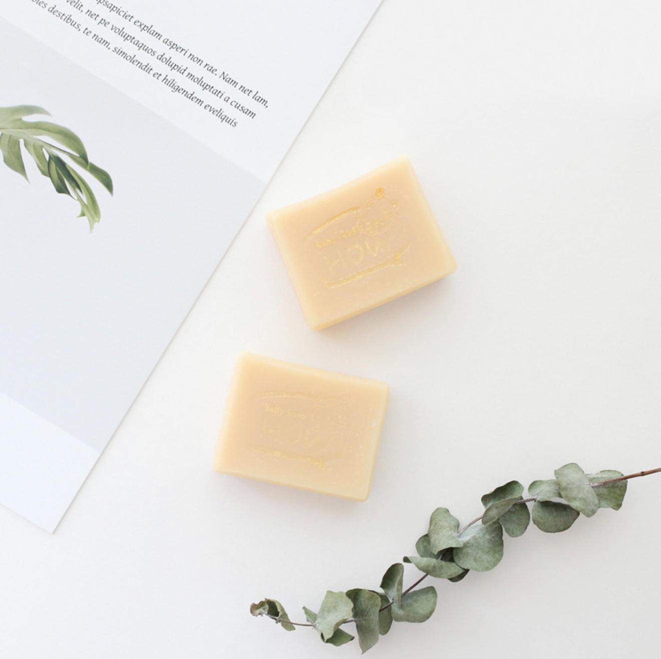 A minimalist presentation of two cream-colored bars of soap resting on a clean white surface, accompanied by a sprig of eucalyptus and a partially visible open book or pamphlet, evoking a sense of natural purity and tranquility.