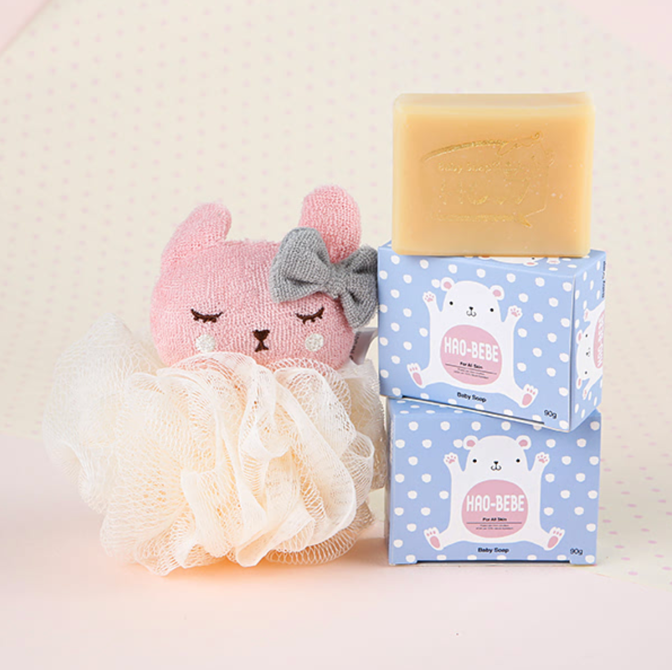 A playful arrangement featuring a stack of "HAO-BEBE" baby soap bars in adorable pastel blue packaging with a cartoon polar bear design, paired with a soft pink plush bunny toy and a delicate cream bath loofah, emphasizing gentle care for children and family.