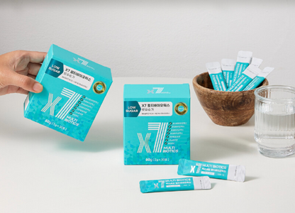 A light teal-colored box of X7 Low Sugar Multi Biotics being held by a hand, with a wooden bowl filled with individual teal sachets and a clear glass of water in the background.