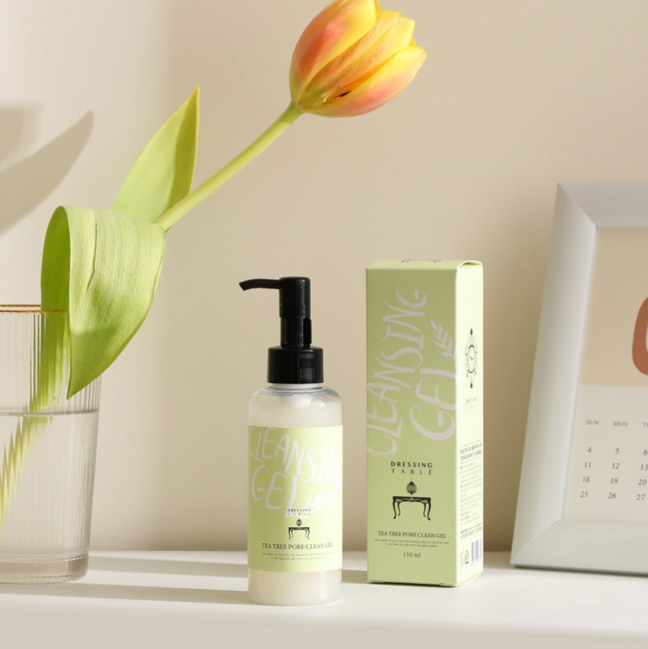 A bottle of [DressingTable] Tea Tree Pore Cleansing Gel placed next to its box and a yellow tulip in a glass vase, styled on a minimalist white table with soft natural lighting.