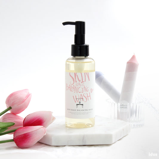 A clear bottle of [DressingTable] Skin Magic Balancing Wash with a sleek black pump, placed on a white marble surface decorated with soft pink tulips and pastel-colored candles.