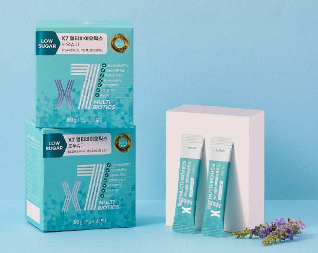 Two teal-colored boxes of X7 Low Sugar Multi Biotics stacked on a light blue background, with two individual sachets leaning against a white block and small lavender flowers for decoration.