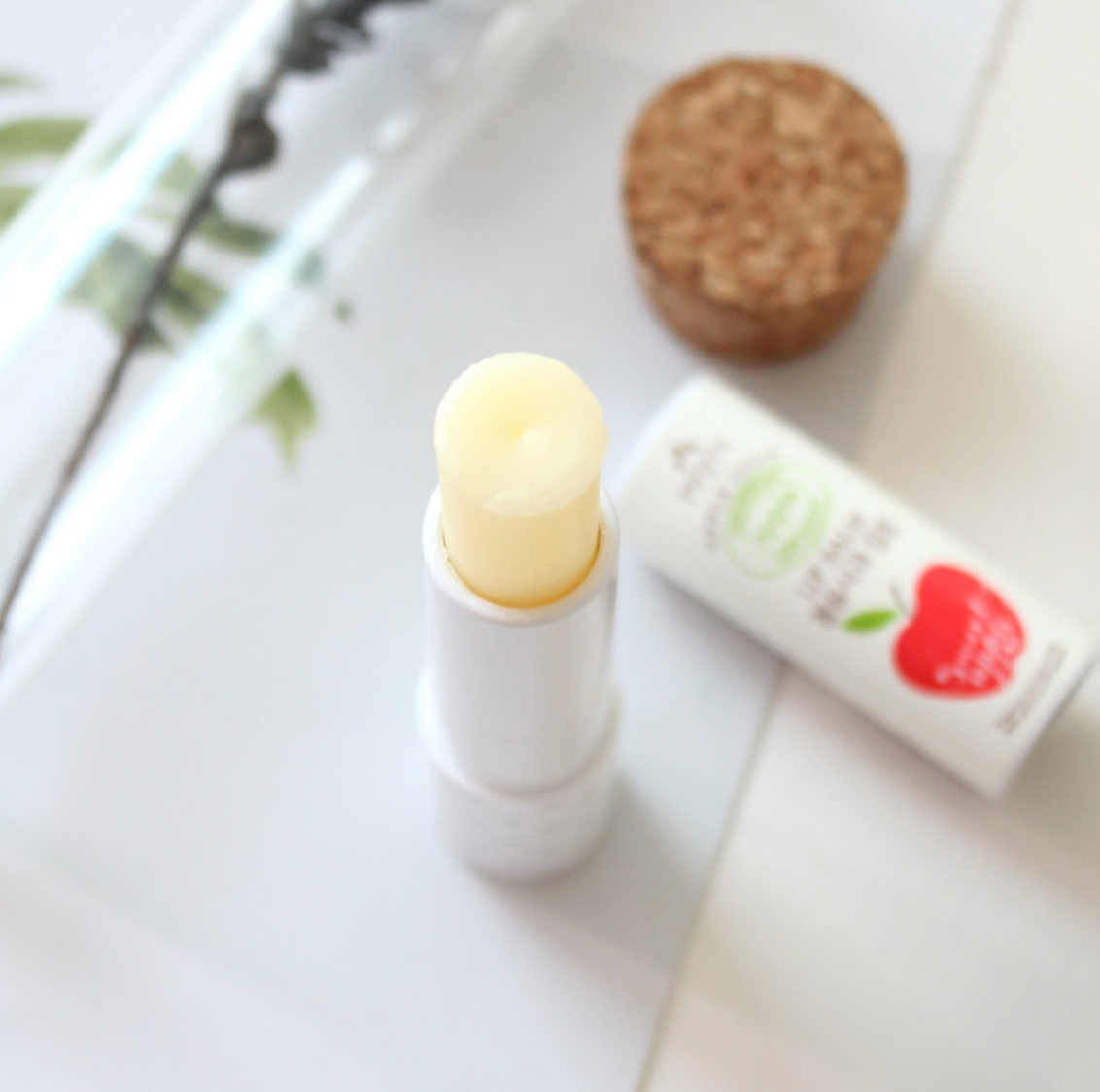 A close-up of the [DressingTable] Apple Lip Balm tube with the balm exposed. The minimalist packaging is shown alongside natural elements, including a cork and decorative glass tube, on a soft background.