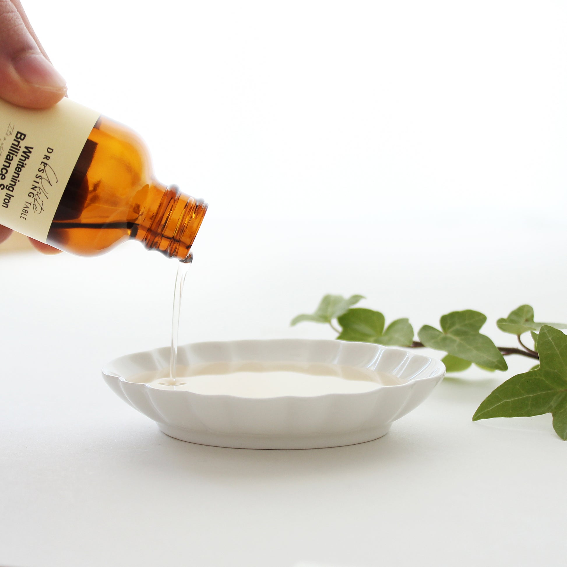 A close-up of Brightening Iron Brilliance Serum being poured into a white dish, showcasing its lightweight, translucent texture. Green ivy leaves add a natural touch to the serene composition.