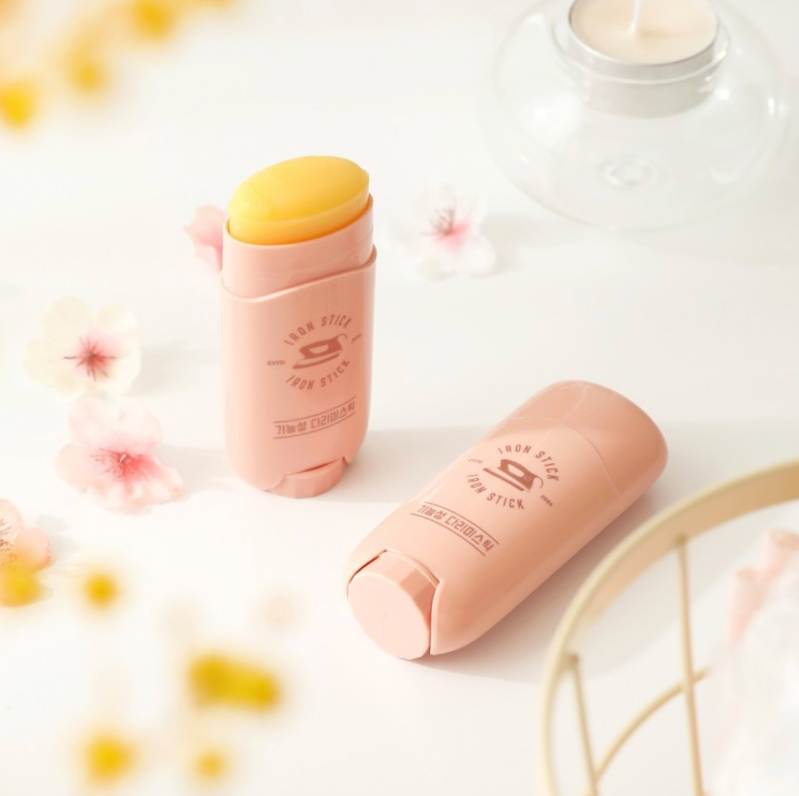 Two open Iron Stick containers with a pale pink design placed on a white table, surrounded by delicate pink flowers, showcasing the product's solid balm texture.