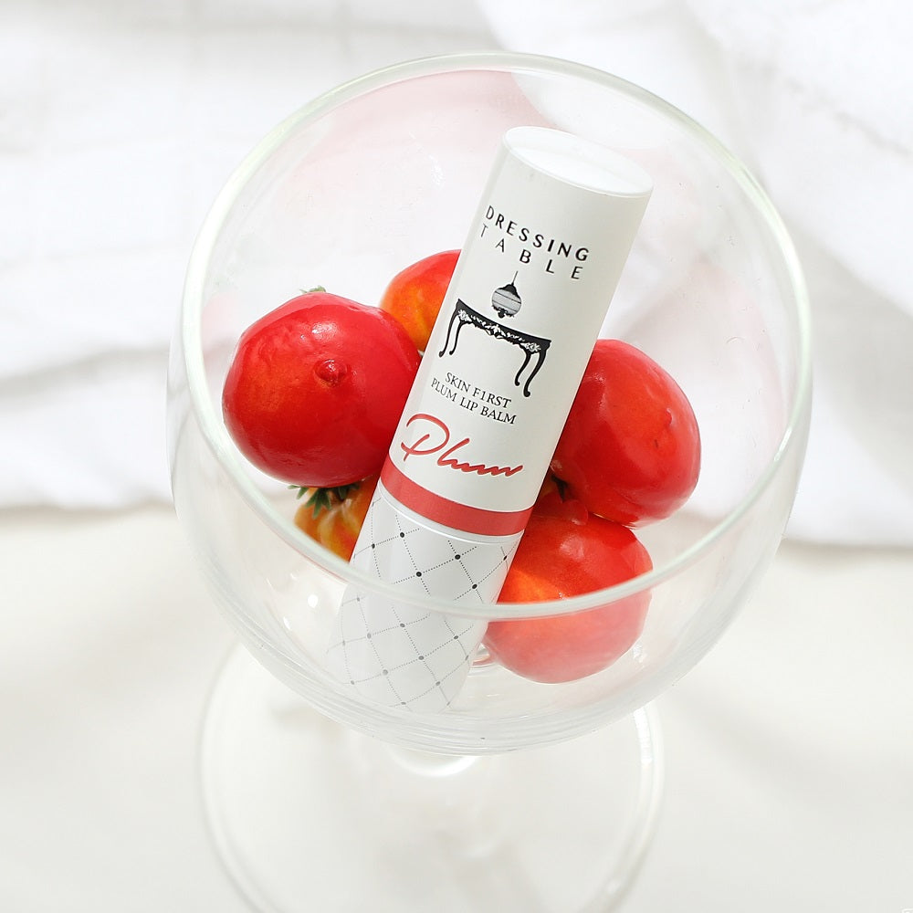 A tube of DressingTable Plum Lip Balm displayed inside a clear glass bowl filled with small red fruits, emphasizing its natural and fresh concept.