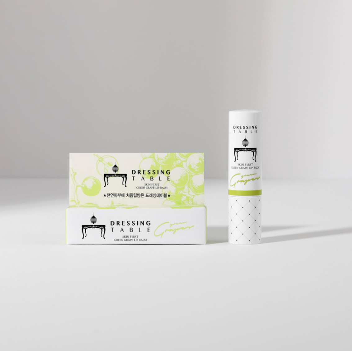 A minimalist display of [DressingTable] Green Grape Lip Balm, with the tube standing upright next to two matching product boxes featuring green grape illustrations.