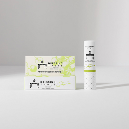 A minimalist display of [DressingTable] Green Grape Lip Balm, with the tube standing upright next to two matching product boxes featuring green grape illustrations.