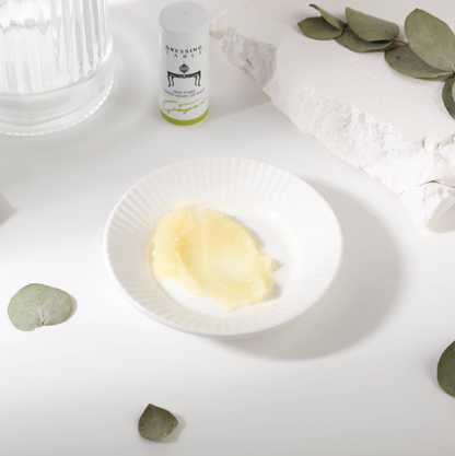 A close-up of a creamy texture of [DressingTable] Green Grape Lip Balm in a white dish, with the product tube and natural eucalyptus leaves arranged on a clean, minimal background.