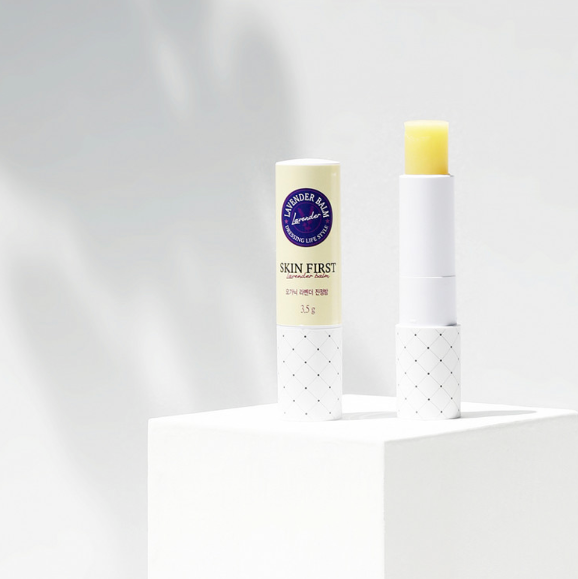 The balm tube, partially open, is showcased on a clean, white pedestal against a bright, minimalistic background. This emphasizes the sleek design and sophistication of the product.