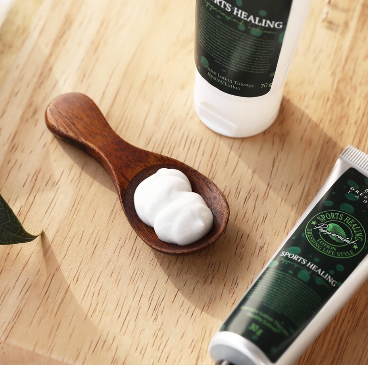 A close-up of a wooden spoon holding a dollop of creamy [DressingTable] Sports Healing Lotion, placed on a wooden surface alongside the product tubes, emphasizing the lotion's smooth texture and natural ingredients.