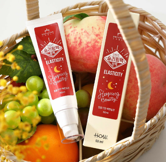 The DressingTable Elasticity Sleeping Beauty Pack tube and its matching box displayed in a woven basket filled with fresh peaches, green grapes, and other fruits, symbolizing the product's natural and nourishing qualities.