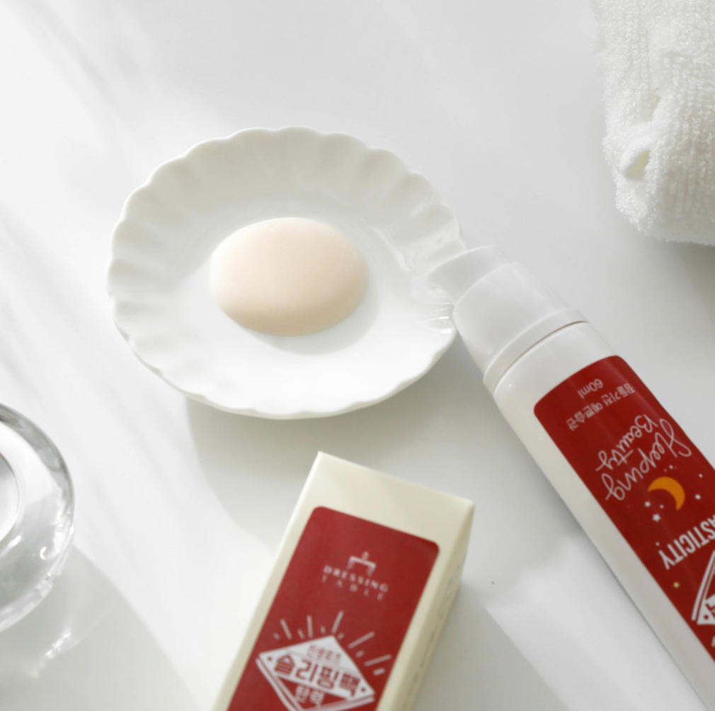 A close-up of a white ceramic plate holding a dollop of the DressingTable Elasticity Sleeping Beauty Pack, showcasing its smooth texture. Next to the plate, the product tube with its red label and a box are placed neatly on a white surface.