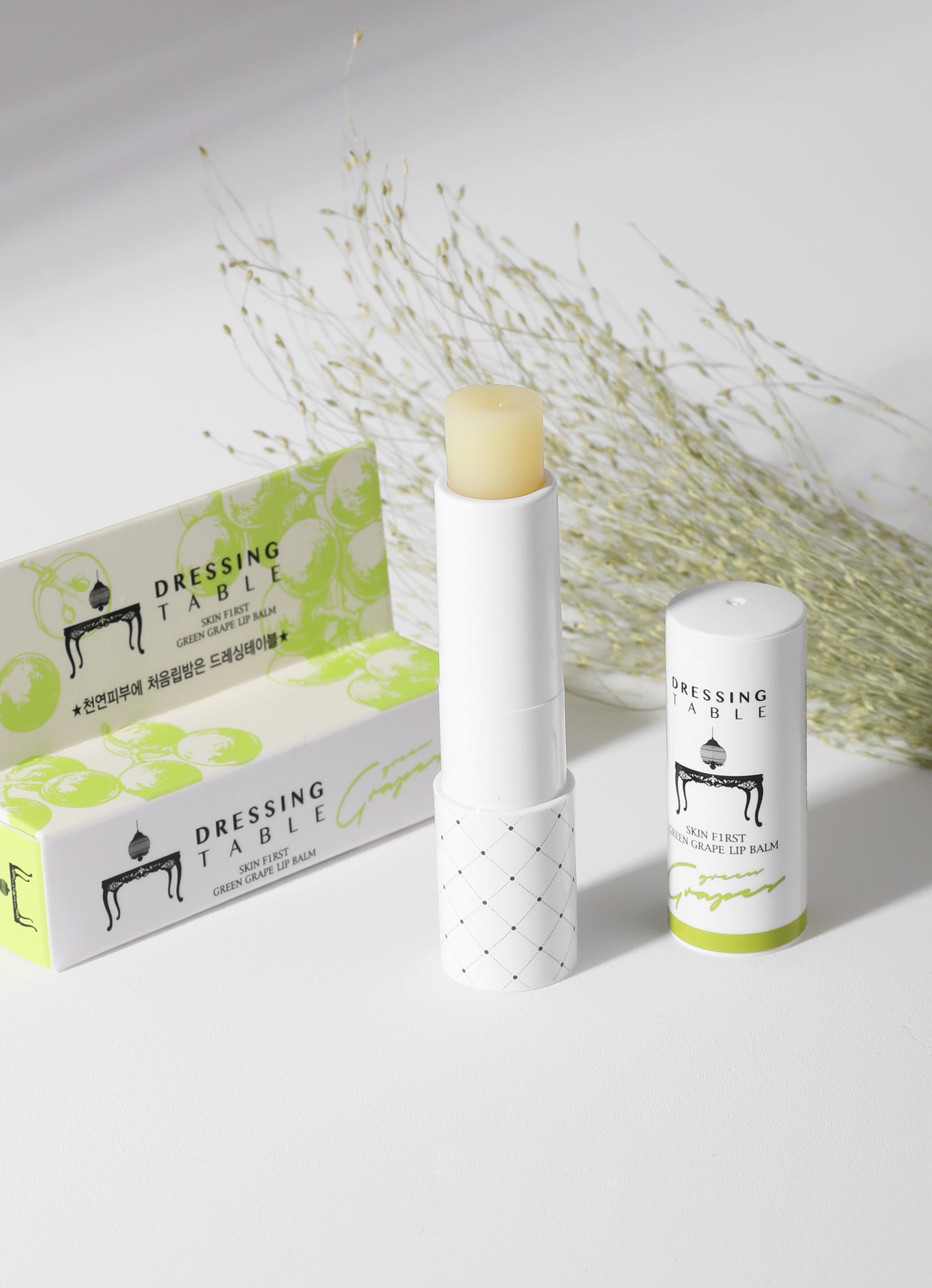 A tube of [DressingTable] Green Grape Lip Balm with its cap removed, revealing the pale yellow balm. The product is displayed next to its green grape-themed packaging, with dried floral elements in the background.