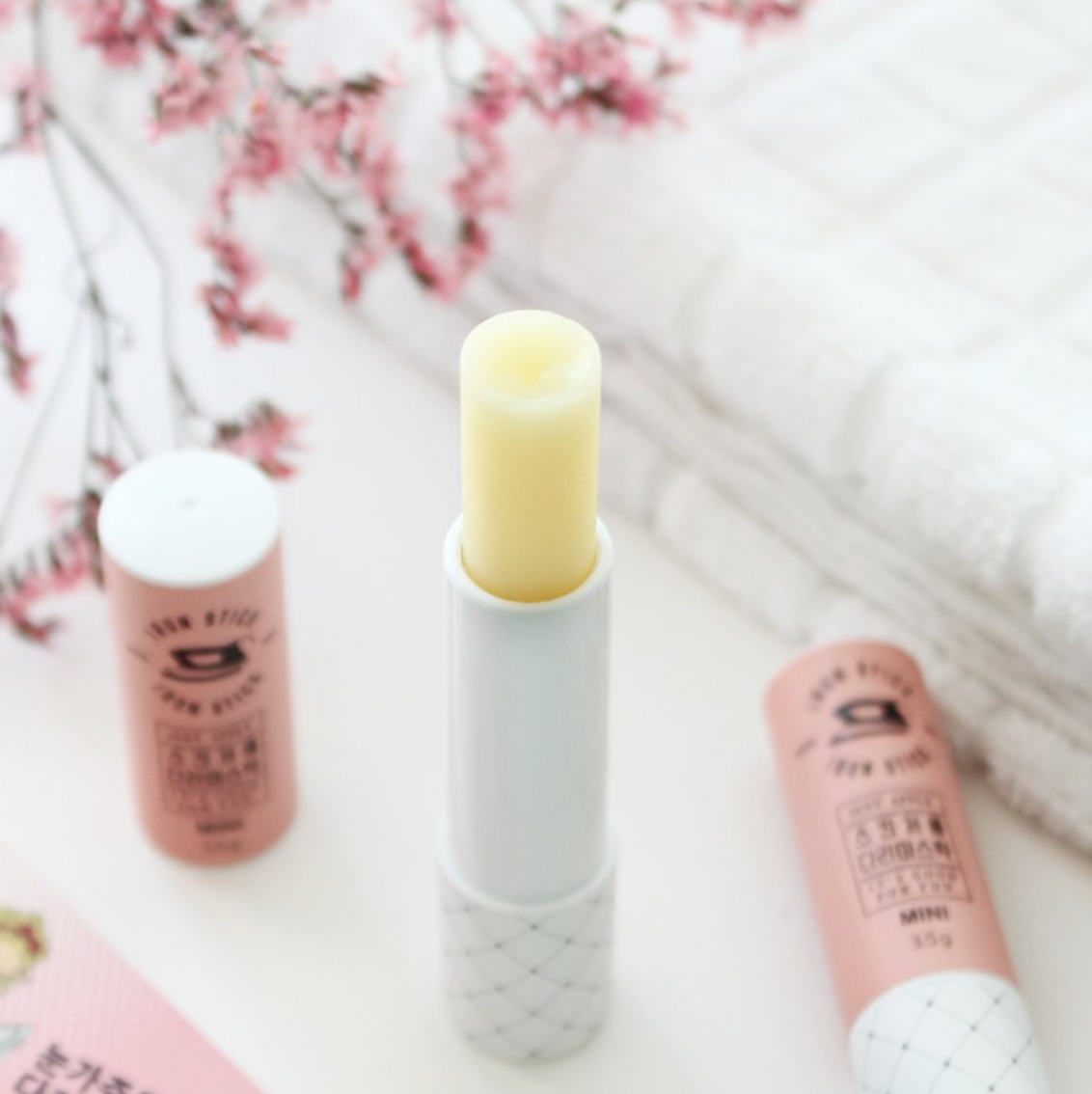 Close-up of an open mini Iron Stick with its creamy balm texture exposed, set against a soft white towel and pink flowers for a clean, fresh aesthetic.